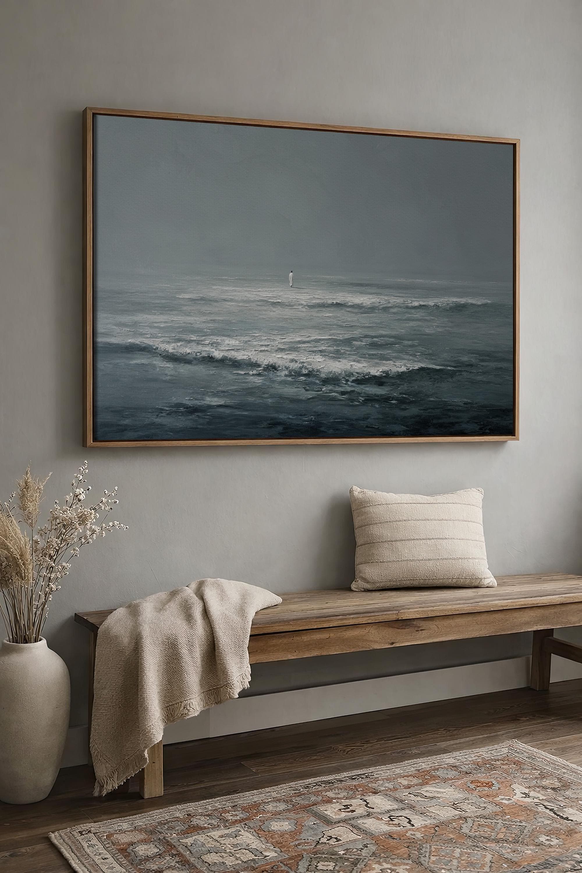 May include: A framed seascape painting depicts a solitary figure standing in the ocean. The artwork, in shades of gray and blue, hangs above a wooden bench with a beige throw and pillow. A vase with dried plants and a patterned rug complete the scene.